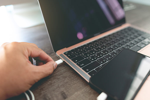 Scene with a person holding a USB Type-C cable connector in their hand and connecting it to a laptop PC. A smartphone with a connector plugged into its USB Type-C port is present in the foreground.