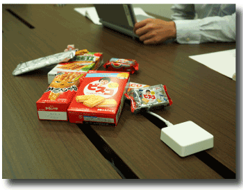 「NAONA」コミュニケーションセンサ（右手前の白い装置）と江崎グリコのお菓子